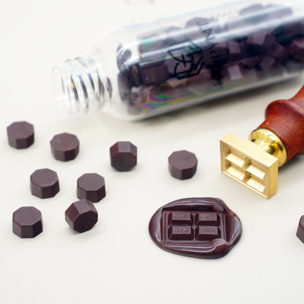Dark brown wax seal beads scattered next to a gold metal wax seal stamp and a finished wax seal.