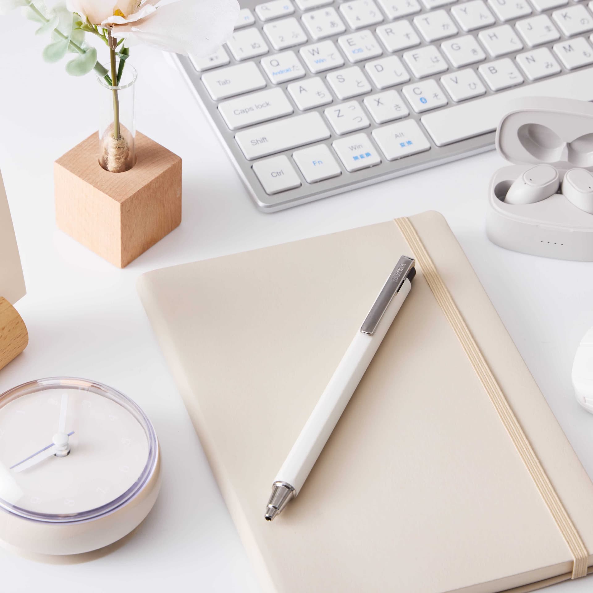 A white retractable ballpoint pen resting on a beige notebook next to a computer keyboard and clock.