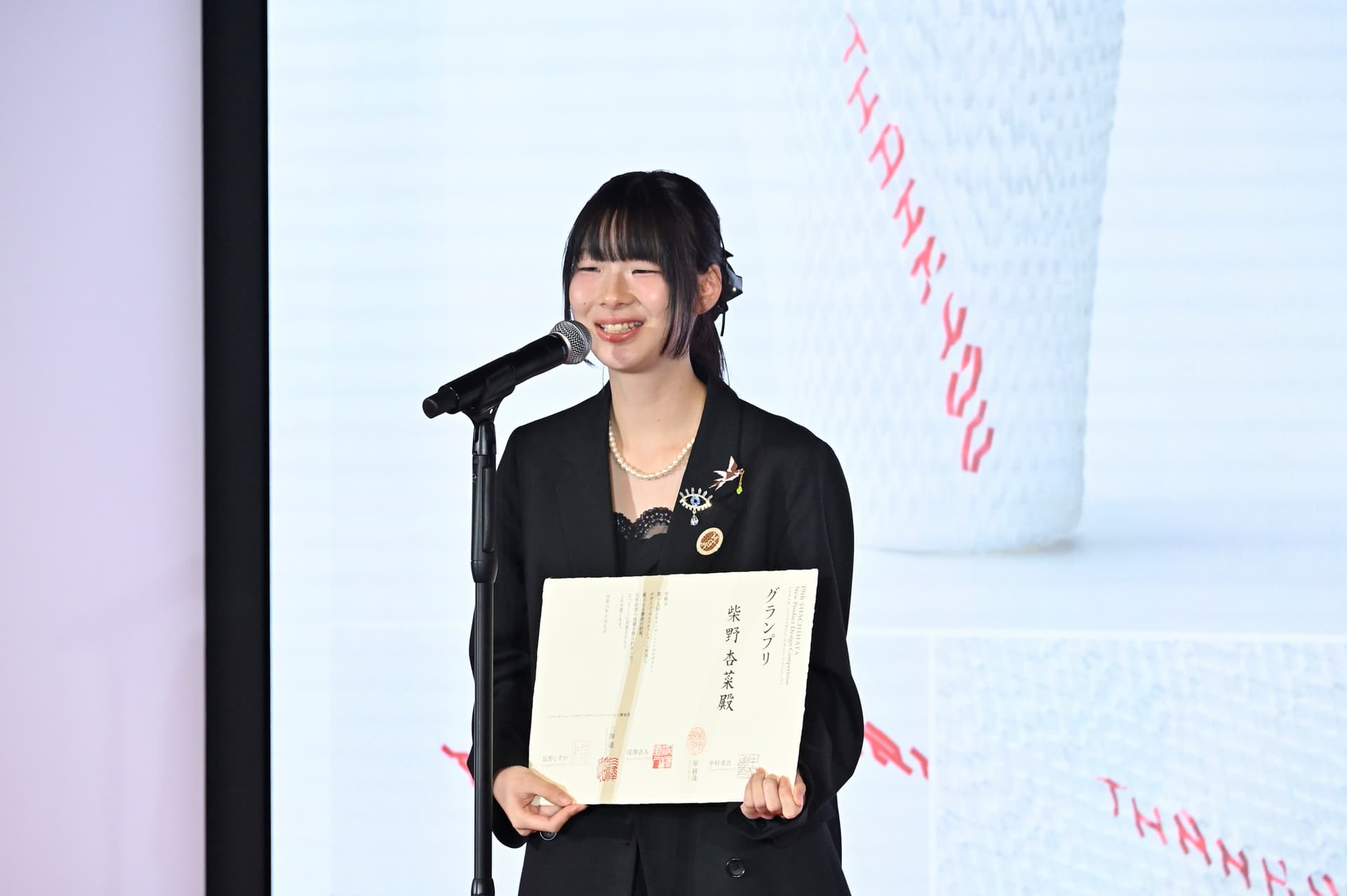 Designer, Anna Shibano, in a black blazer holding a white award certificate in front of a large screen.