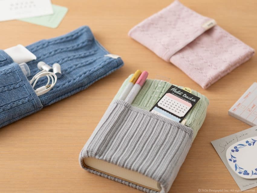 A grey knit fabric notebook cover holding a habit tracker and two pens on a wooden surface.