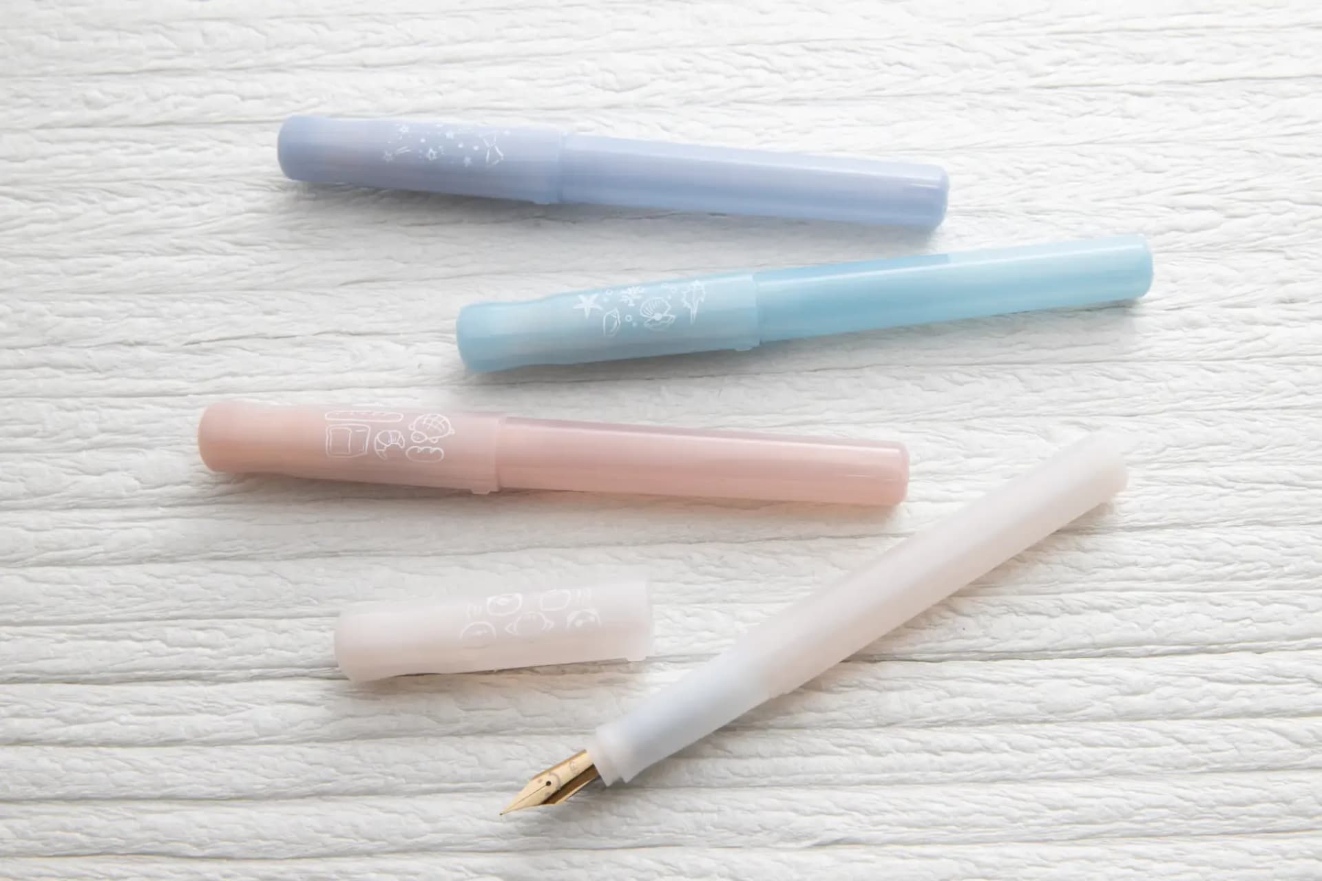 Four Pilot Kakuno Madoromi fountain pens in translucent pastel blue, pink, and white colors arranged on textured paper.