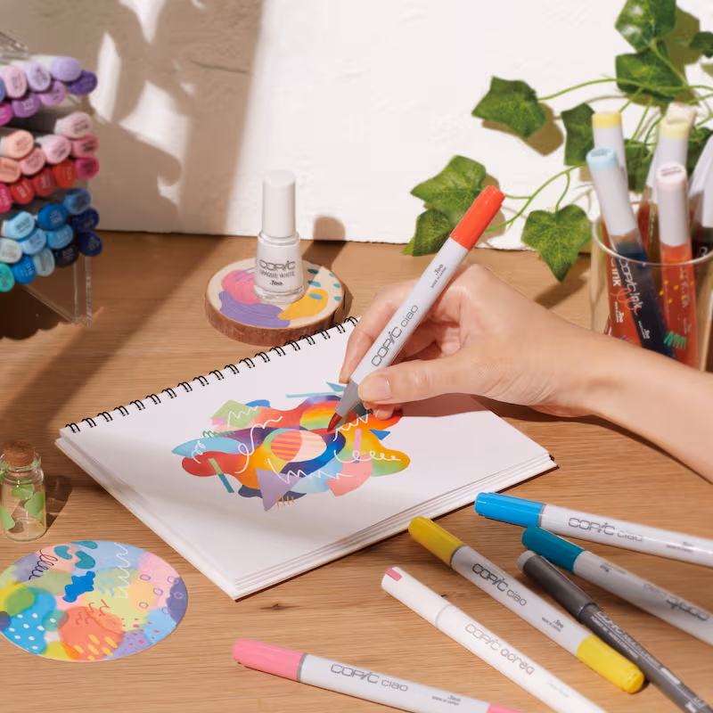 A hand uses a Copic Ciao marker to draw colorful abstract shapes in a spiral notebook.