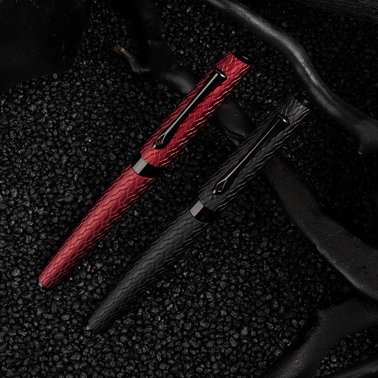 Two textured metallic rollerball pens in deep red and matte black resting on dark volcanic stones.