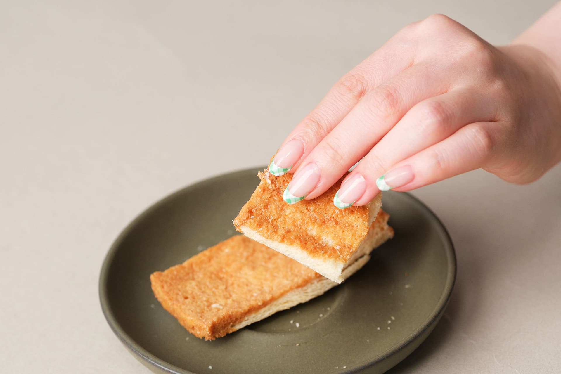 A hand with white manicured nails picks up a piece of golden brown toasted bread from a dark plate.