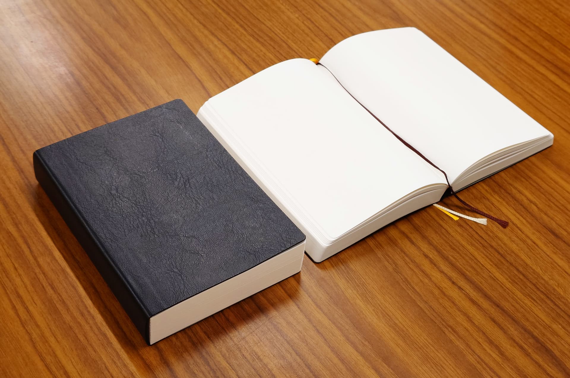 Closed and open black textured hardcover notebooks with white pages and ribbon bookmarks on a wooden surface.